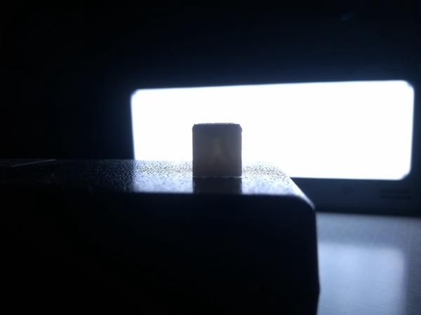 A 3D-printed calibration cube, posed in front of a light source for better visibility of its problematic features. Its first few layers show a taper, starting small, before increasing to the expected size. One size shows a bulge, where layers are too large.