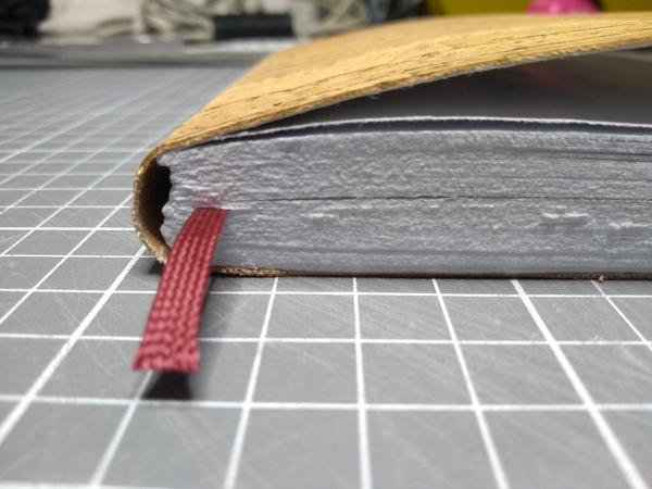 A closeup of bottom side of the notebook, the bookmark sticking out. The edges of the paper and cover are visibly rough.