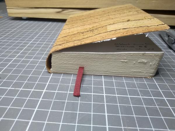 A closeup of bottom side of the notebook, the bookmark sticking out. The edges of the paper and cover are visibly rough.